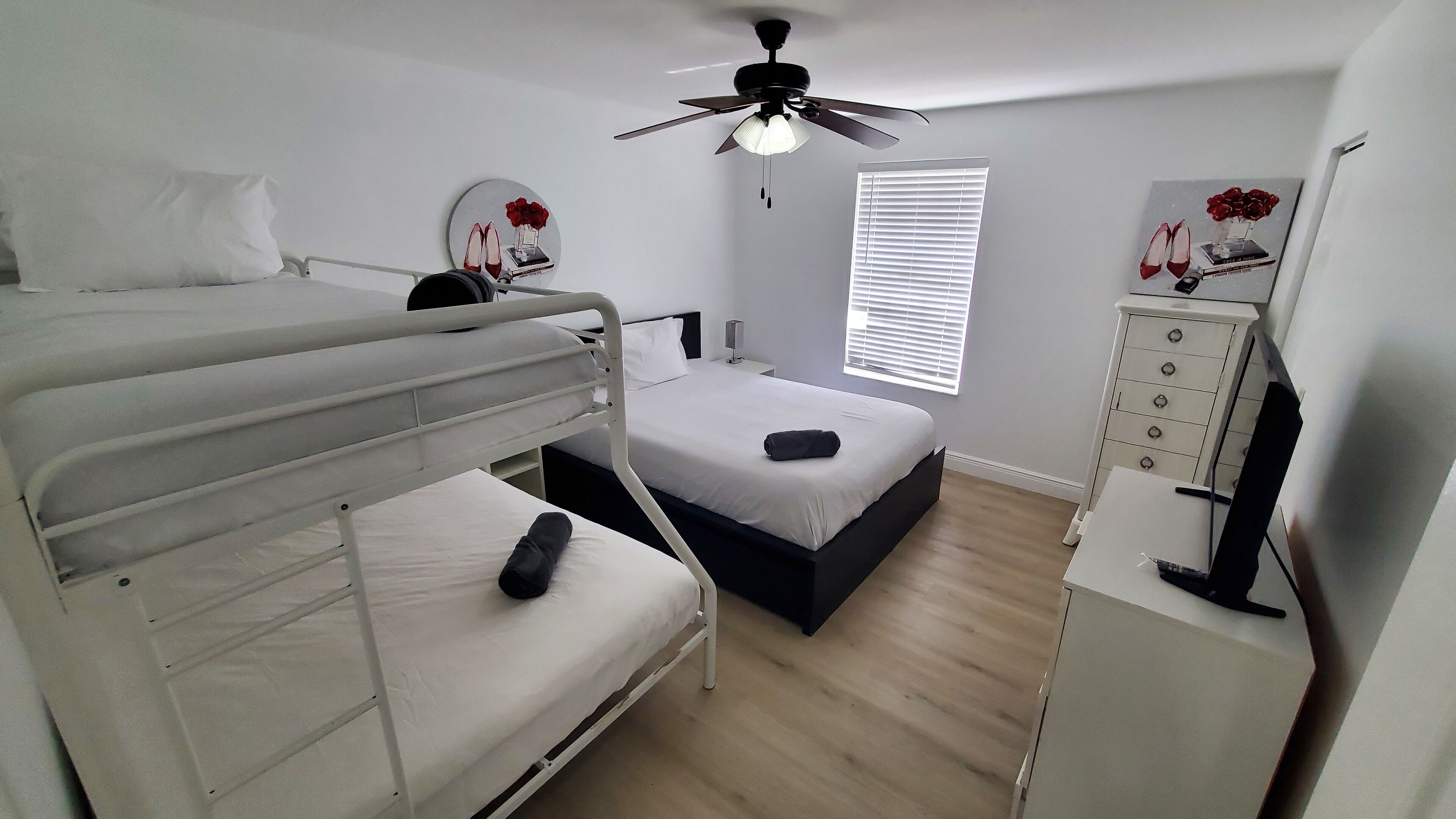 Ein Schlafraum mit Etagenbetten, einem Deckenventilator, einem Nachttisch mit Lampe, einem Fenster mit Jalousien und einer Kommode mit Schubladen.