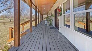 A covered porch with wooden railings, potted plants, and large windows offering views of a park. - Welcome to our picturesque ranch oasis less than 1 hour from DFW!  (Cleburne)