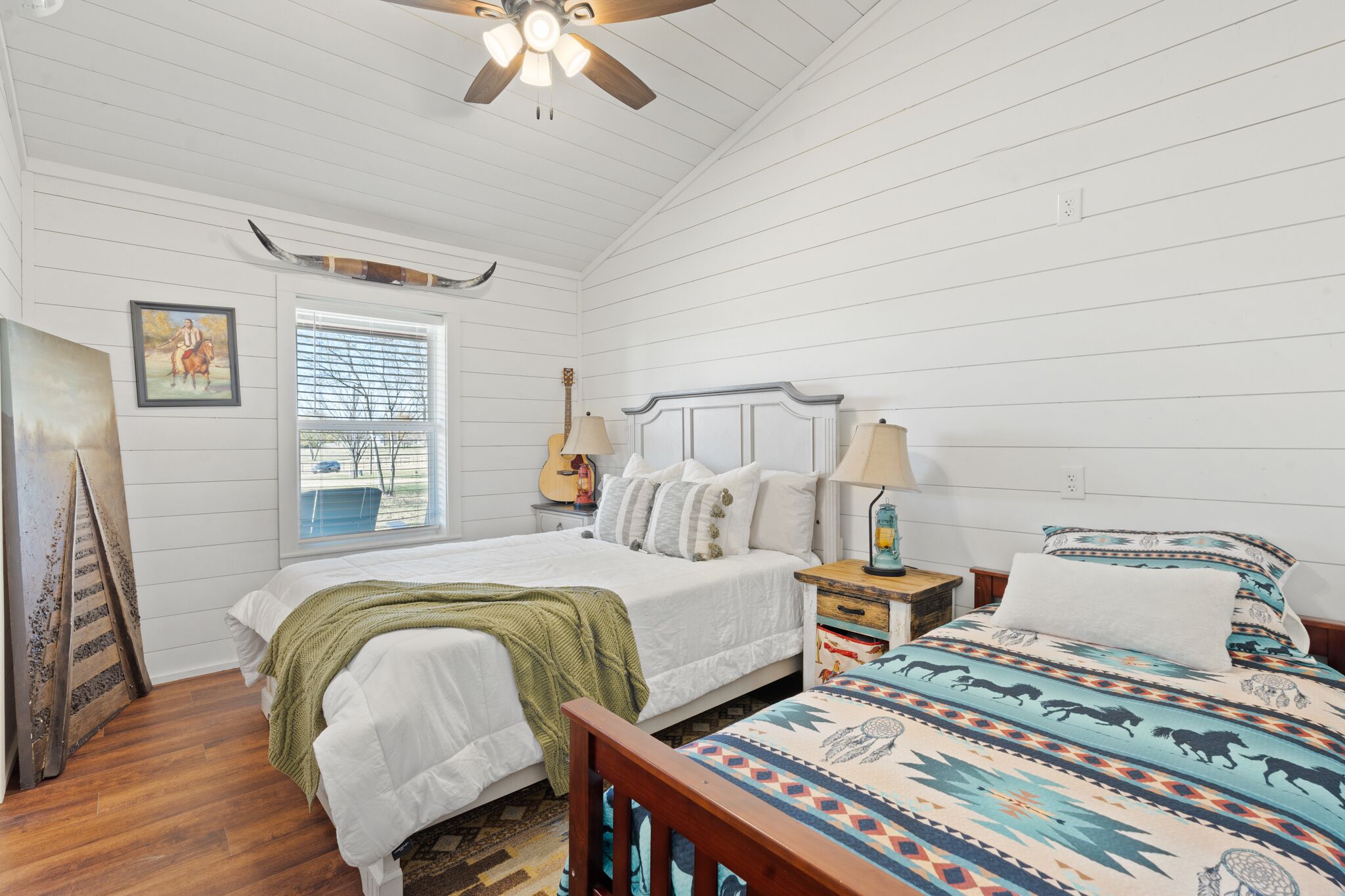 A bedroom with a wooden ceiling, a bed with a green blanket, a patterned bedspread, a guitar, and a window with blinds.