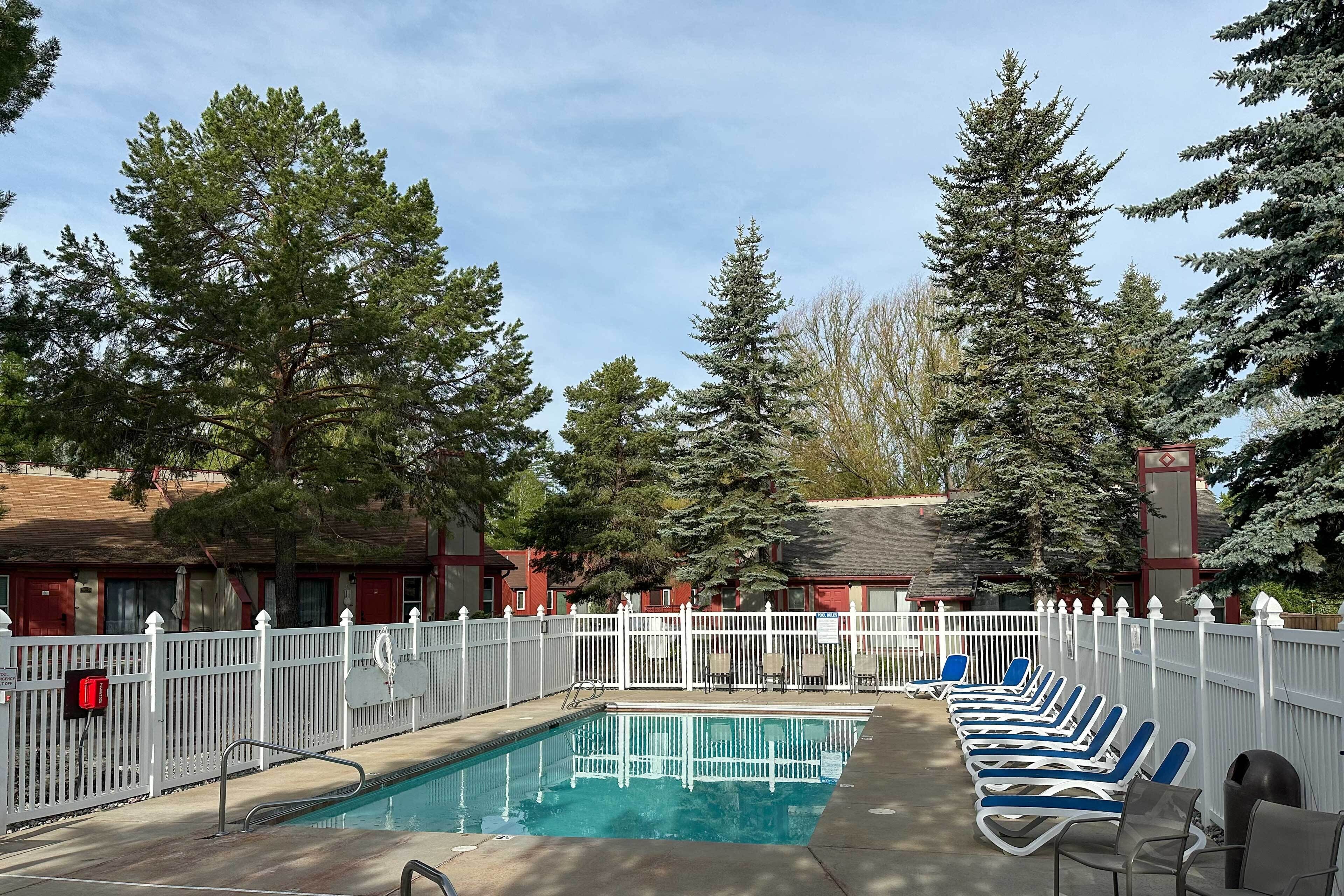 Zona de piscina con valla blanca, sillas reclinables azules y árboles al fondo.