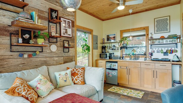 A cozy living room with a sofa, a coffee table, and a red throw blanket. There is a kitchen area with a microwave, a coffee maker, and a refrigerator. - Yadkin River 'treehouse'~ Miles of Trails On-site (East Bend)