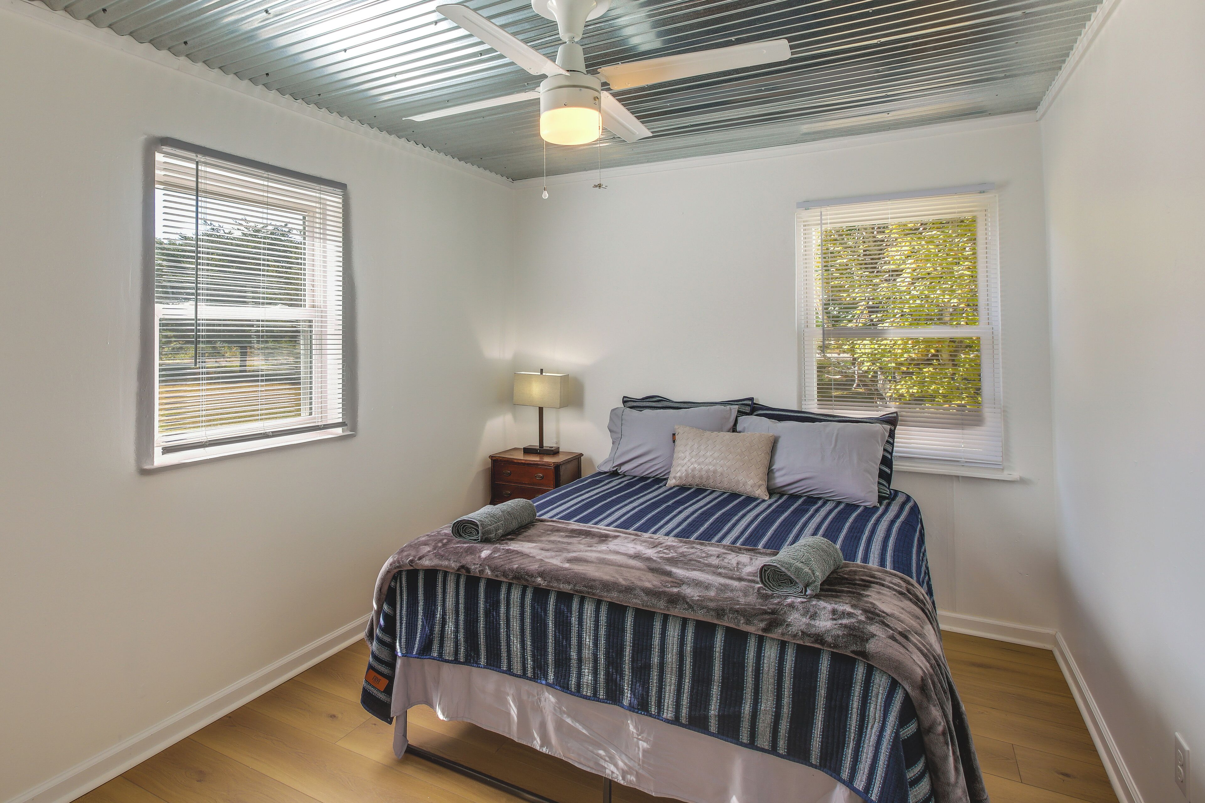 A bedroom with a bed, two windows, a nightstand, a lamp, and a ceiling fan.