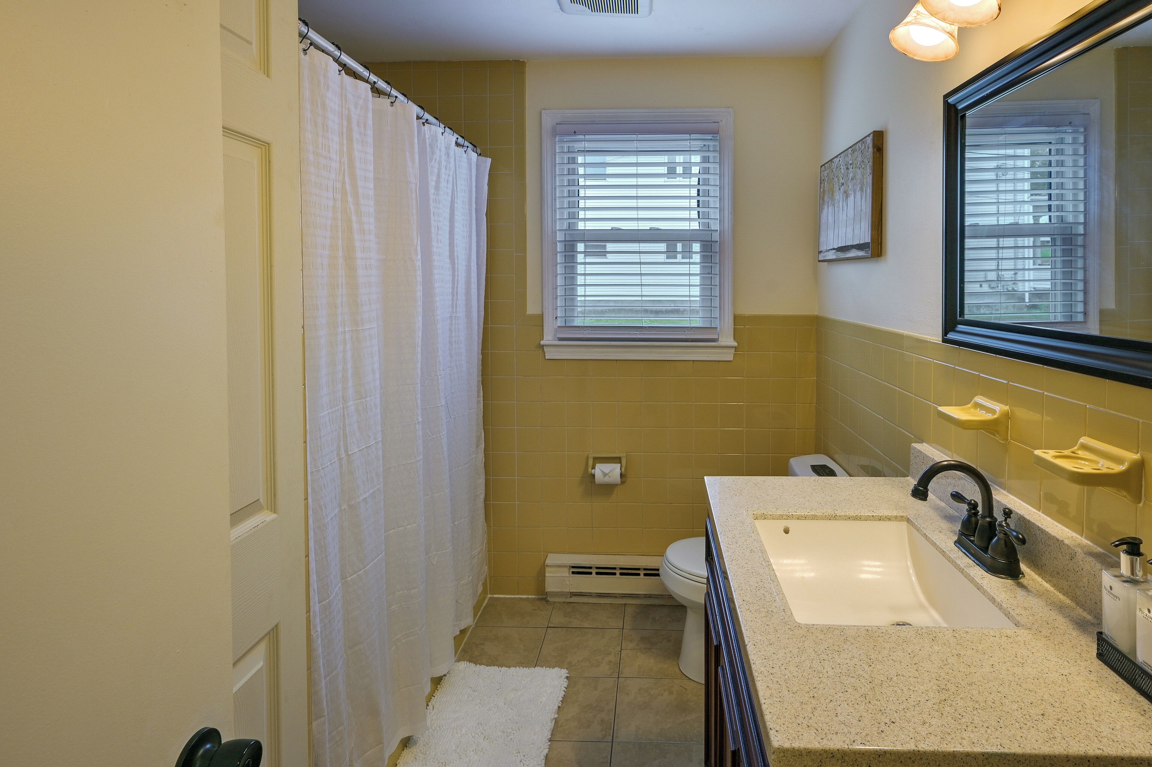 Une salle de bain avec un lavabo blanc, un robinet noir, un rideau de douche blanc et une fenêtre avec des stores.