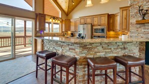 A spacious kitchen with wooden cabinets, a large island with stools, a stone backsplash, and a view through the sliding glass doors. - Hot Tub & Fire Pit: Idyllic Getaway in Angel Fire! (Angel Fire)