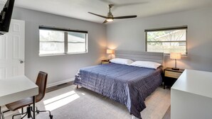 Une chambre à coucher comprenant un lit, deux lampes de chevet, un ventilateur de plafond, un bureau avec une chaise et deux fenêtres.
