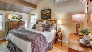 A bedroom with a wooden ceiling, a bed with multiple pillows, a nightstand, a lamp, and a window with a view of trees.