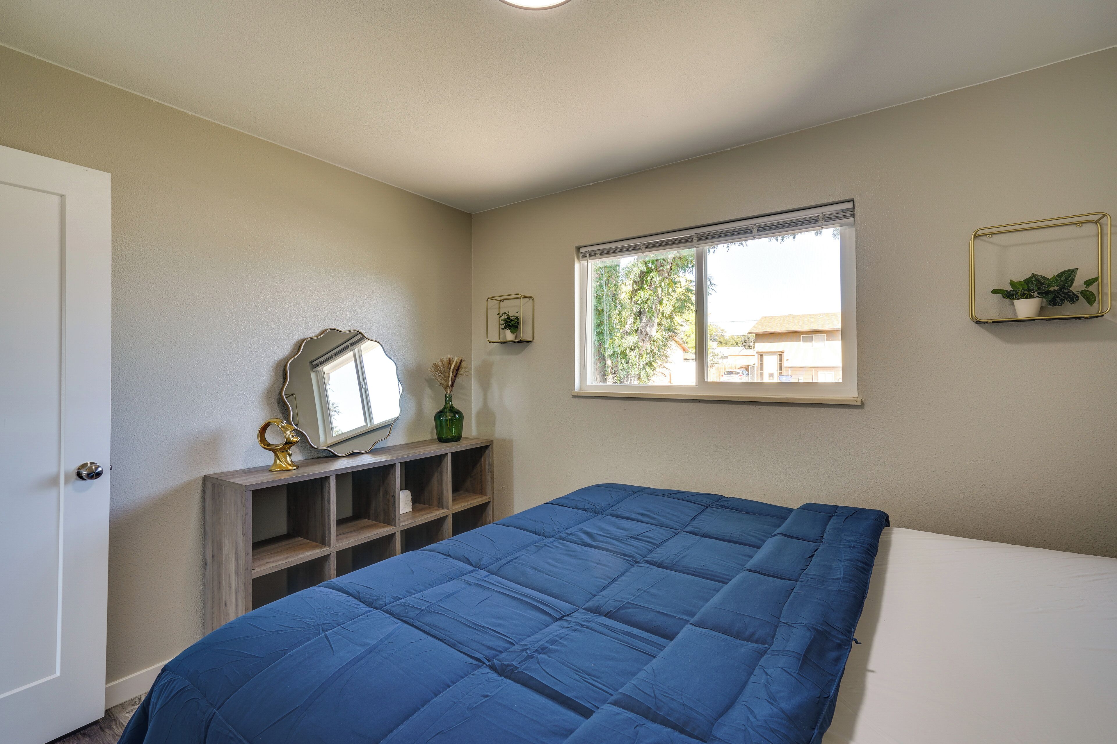 Une chambre avec un grand lit, une fenêtre avec des stores, un miroir et une petite étagère sur laquelle est posée une plante.