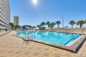 A swimming pool with a tiled deck, surrounded by palm trees and a tall building in the background.