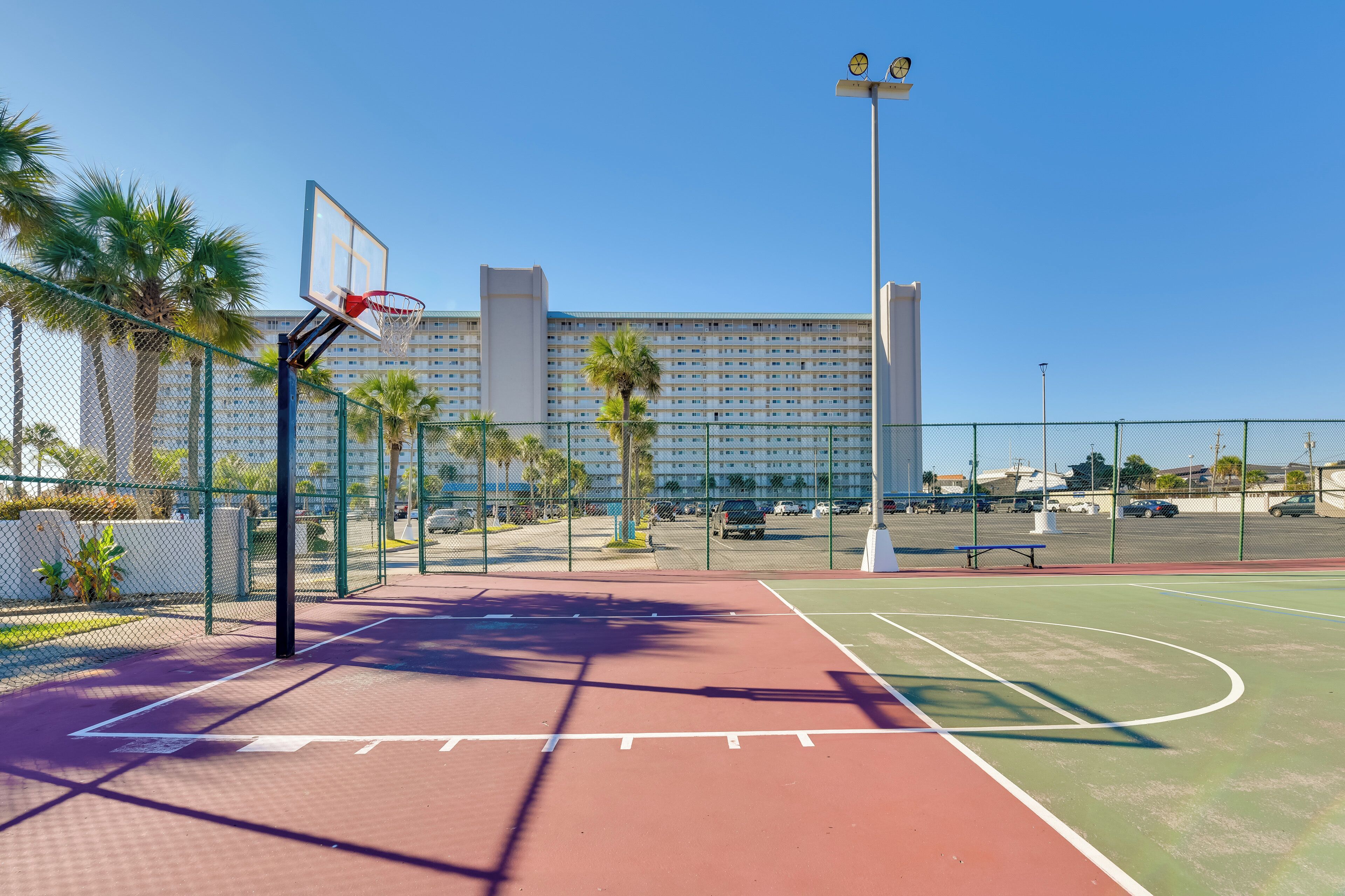 Een buitenbasketbalveld met een hek van gaas, palmbomen en een groot gebouw op de achtergrond.
