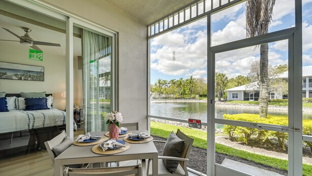 A room with a dining table, chairs, a bed, a ceiling fan, and a large window with a view of a lake and palm trees. - Newly Updated Golf Villa w/ Free Golf May-oct (Naples)