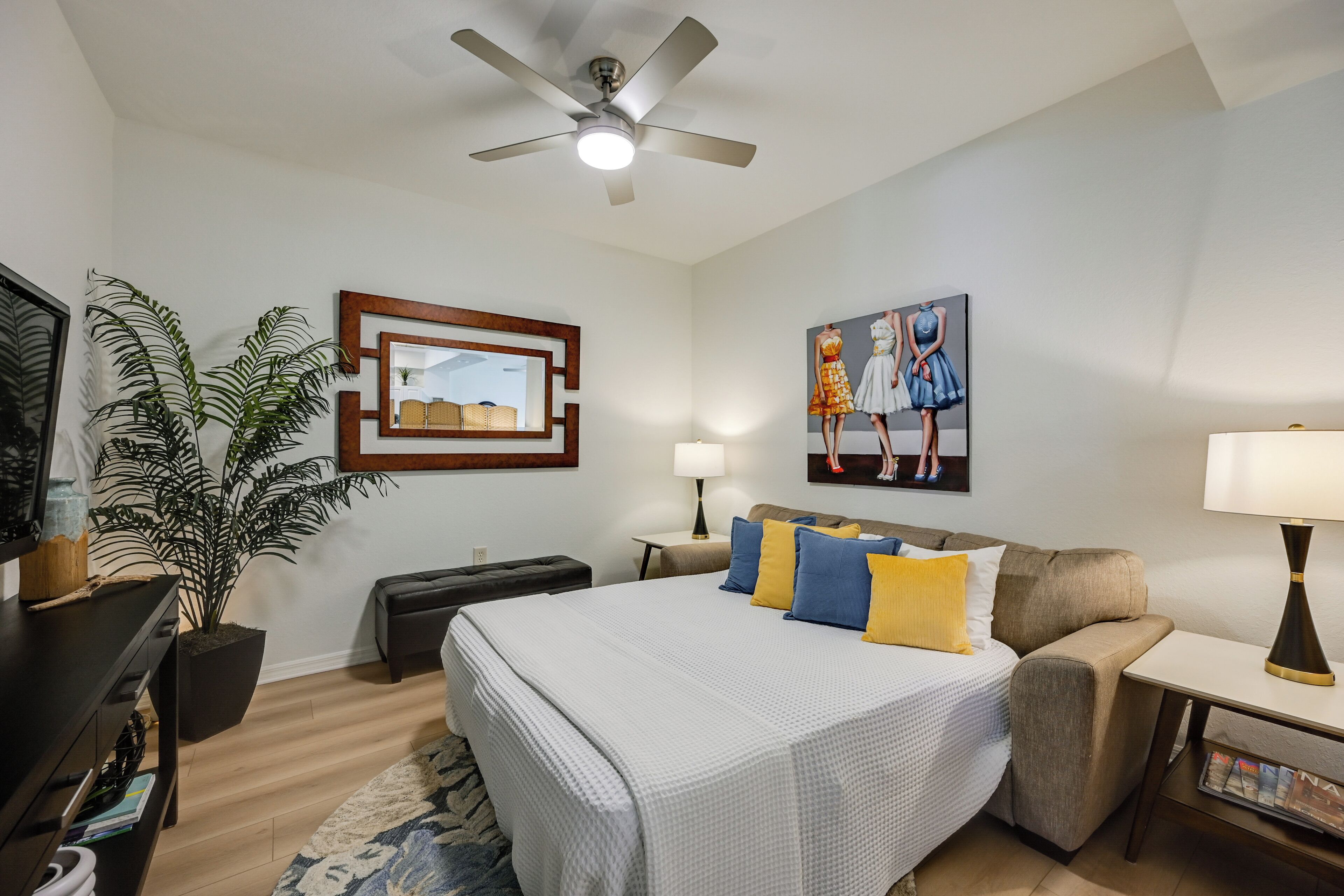 A hotel room with a bed, a desk, a chair, a ceiling fan, a potted plant, a painting, and a lamp.