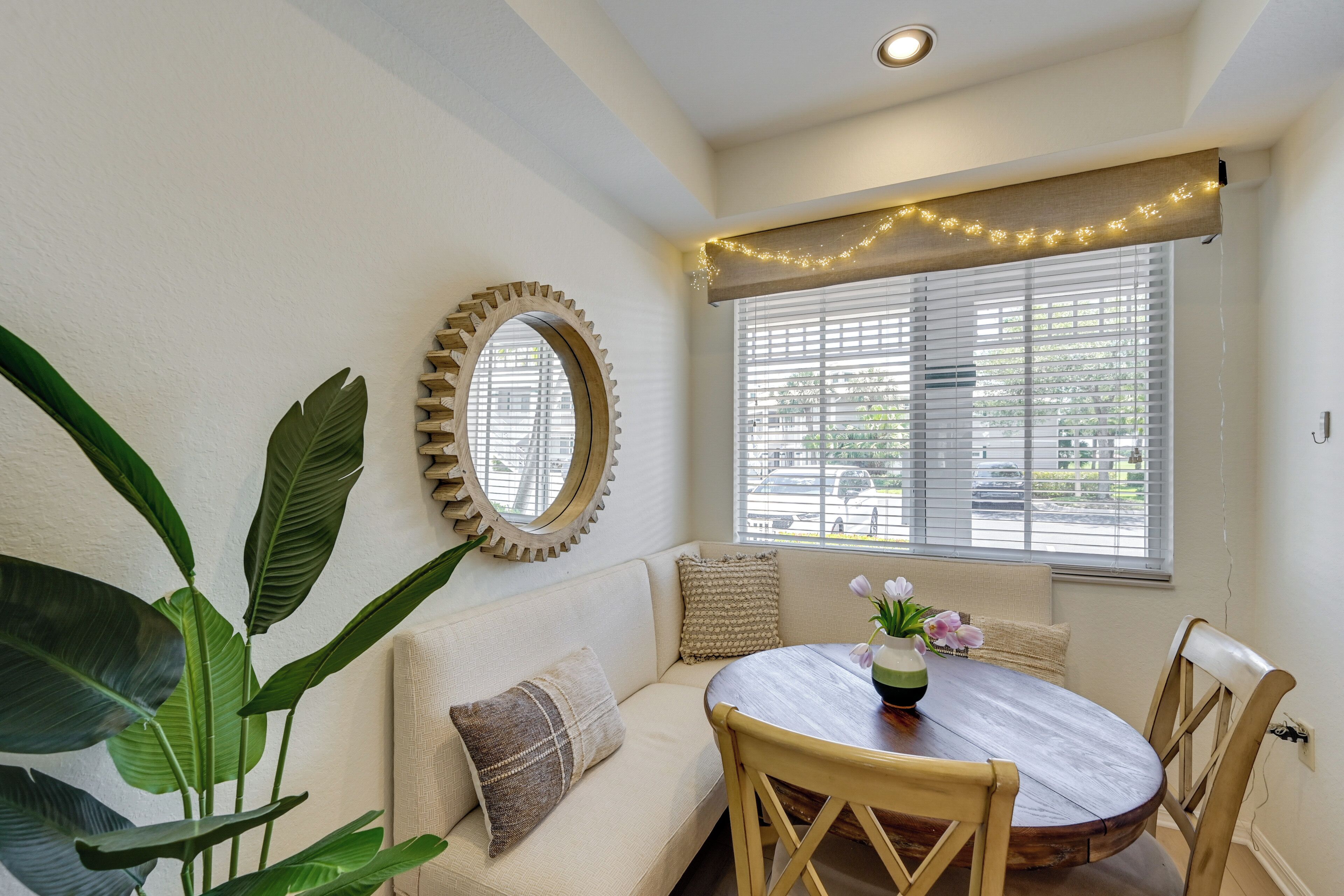 A room with a round wooden table, beige sofa, and a large window with blinds.