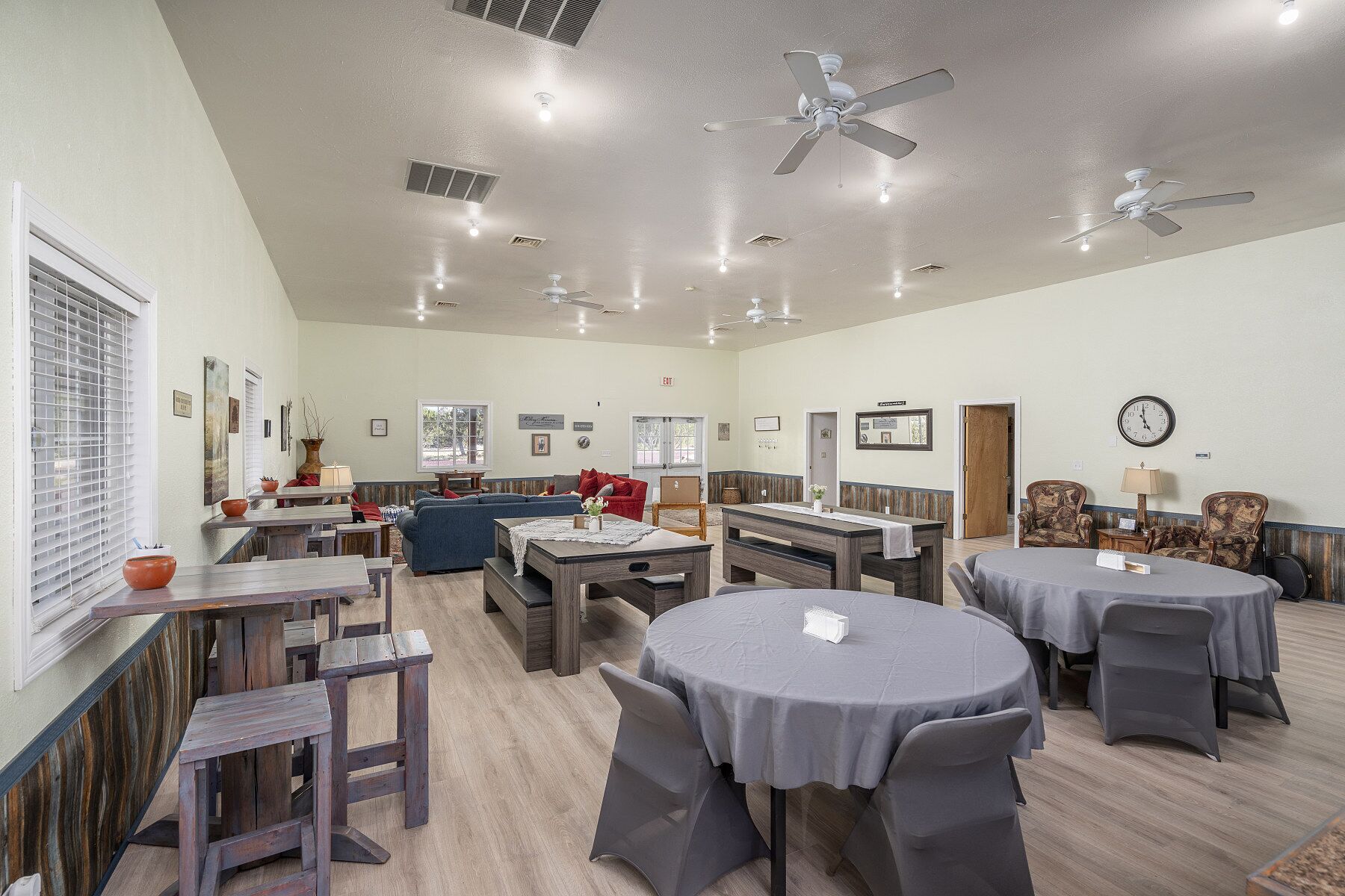 Une pièce spacieuse, dotée de plusieurs tables et chaises, de ventilateurs de plafond et d’une horloge au mur.