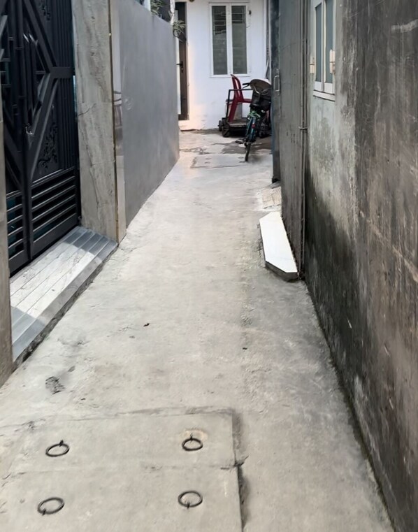 A narrow concrete pathway between buildings with a metal gate on the left and a red chair in the background.
