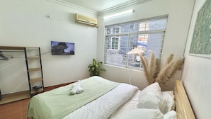 A neatly arranged bedroom with a bed, a TV mounted on the wall, a window with a view of buildings, and an air conditioner.