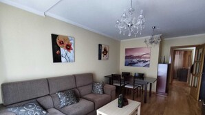 Una sala de estar con un sofá gris, una mesa de centro blanca y un comedor con mesa y sillas. Hay dos cuadros en la pared y una araña.