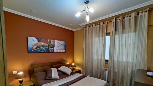 Un dormitorio con cama, dos mesitas de noche con lámparas, una lámpara de techo, un ventanal con cortinas y un cuadro colgado en la pared.
