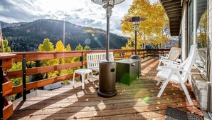 Una terraza de madera con mobiliario exterior, un estufa de leña y vistas a una cadena montañosa boscosa.