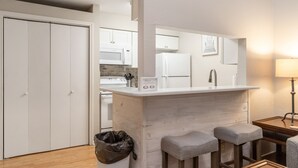 A compact kitchen with white cabinets, a microwave, and a refrigerator. There are two stools and a trash bin. - Cozy Studio Loft | Indoor Pool & Walk to Beach (Corpus Christi)