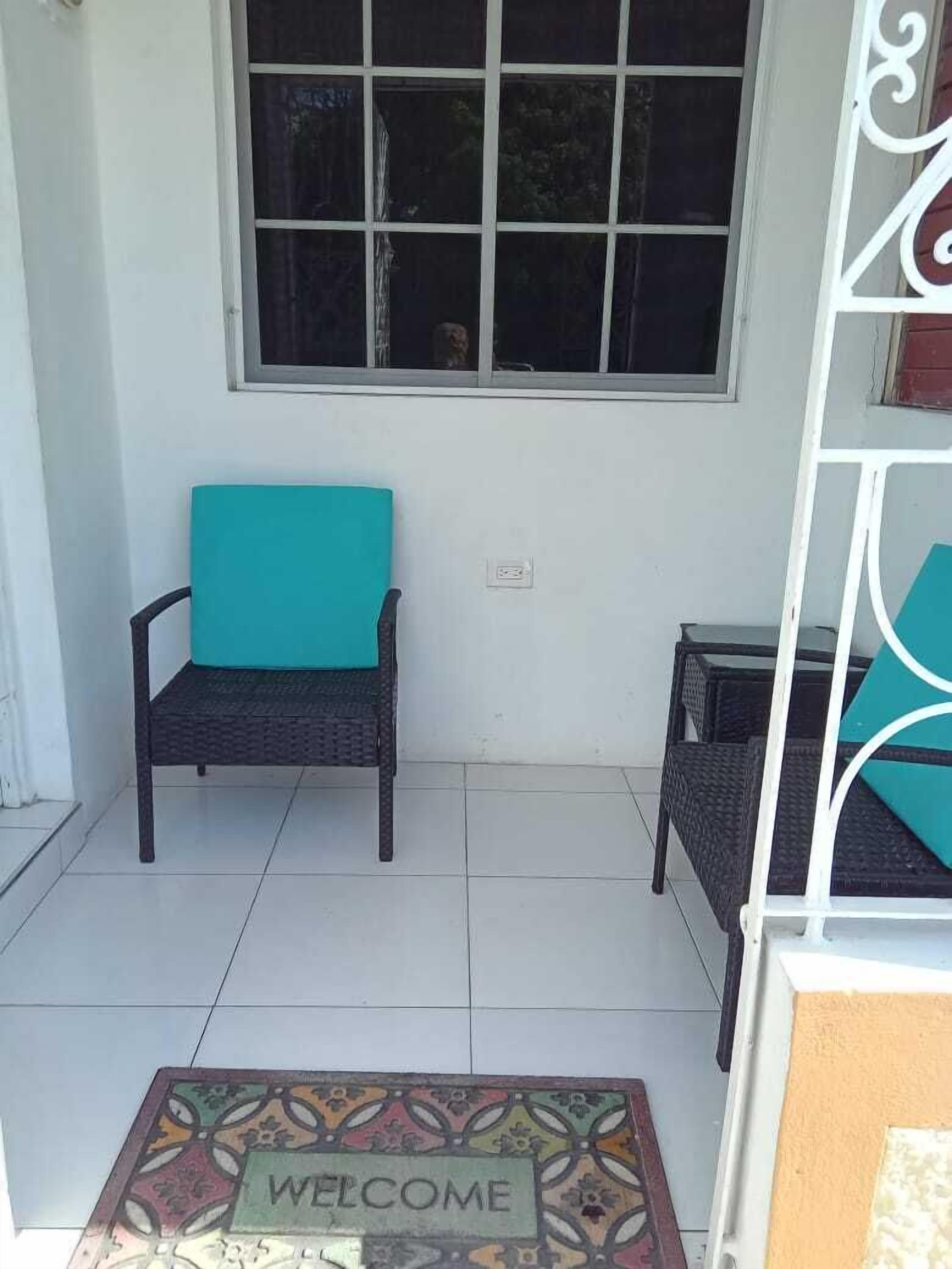 Un pequeño espacio al aire libre con dos sillas de mimbre, un felpudo y una ventana con vistas al exterior.