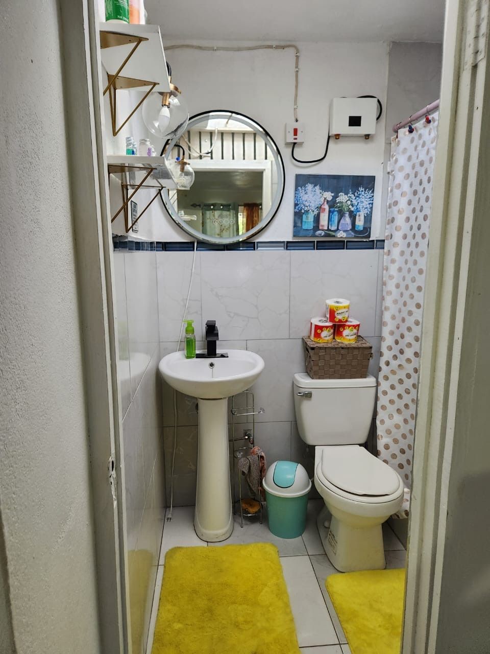A small bathroom with a white sink, a toilet, and a mirror. There are yellow rugs on the floor, and a shelf with toiletries above the sink.