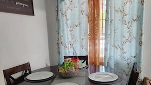 A dining area with a round table set for four, surrounded by chairs with patterned upholstery. A bowl of snacks is placed on the table, and a decorative sign with a biblical verse is visible on the wall.