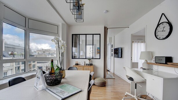 A modern hotel room with a large window, a dining area with a table set for two, a white desk with a computer, and a wall-mounted clock. - Appartement de Charme Hypercentre Brest (Brest)