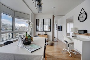 Una habitación de hotel moderna con un ventanal grande, un rincón de comedor con una mesa para dos, un escritorio blanco con una computadora y un reloj de pared.