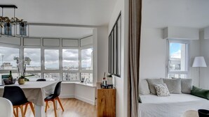 A modern, well-lit room with a large window, a dining area with a table and chairs, a cozy seating area with a sofa and cushions, and a small round table with a bowl. - Appartement de Charme Hypercentre Brest (Brest)