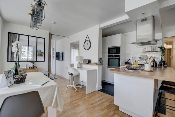 Una cocina moderna con armarios blancos, encimera de madera y horno empotrado. Hay una zona de comedor con mesa y sillas, y un pequeño espacio de oficina con escritorio y silla.