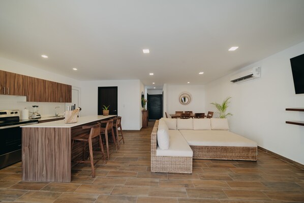 A modern kitchen with a central island, dining area with chairs, a sofa, and a TV mounted on the wall. - Villas Oazis (Taxisco)