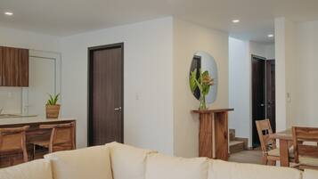 A modern living room with a sofa, wooden chairs, a dining table, a kitchen area, and a large mirror on the wall.