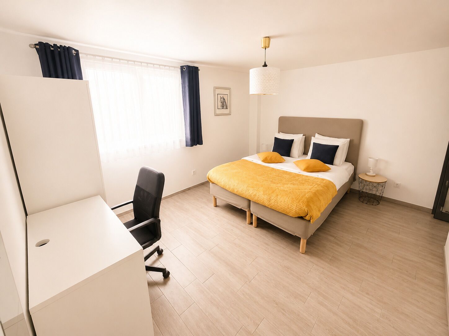 Ett hotellrum med en säng, ett skrivbord, en stol, en lampa och en tavla på väggen.