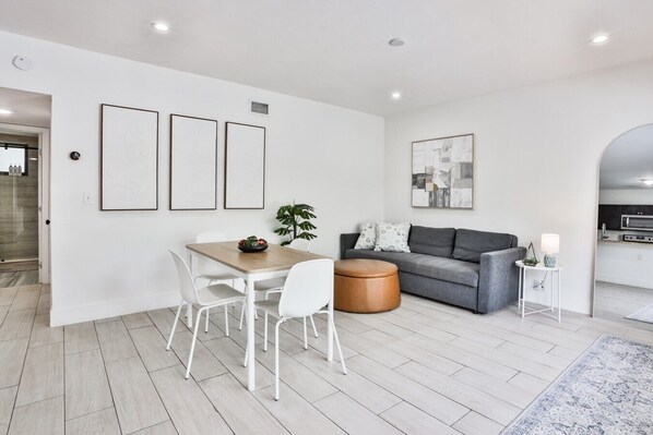 A modern living room with a dining area, grey sofa, and a kitchen visible through an archway.