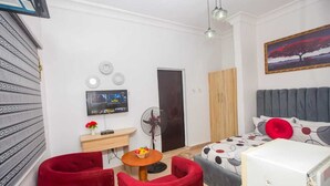 Kompakti hotellihuone, jossa on sänky, televisio, tuuletin, pieni pöytä, jossa on hedelmiä, ja punaiset tuolit.