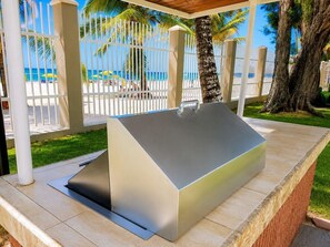Zona de parrilla exterior con mesa metálica y extractor integrado, rodeada por una cerca blanca y palmeras, con vista a una playa.