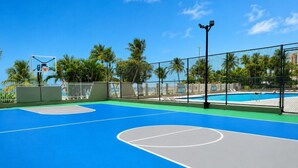 Una cancha de baloncesto al aire libre con superficie azul, rodeada por una cerca, palmeras y una piscina al fondo.
