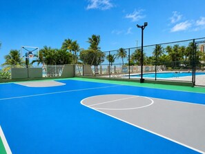 An outdoor basketball court with a blue surface, surrounded by a fence, palm trees, and a pool in the background. - Beachfront Gem Isla Verde Walk 2 Eat W/D (Carolina)