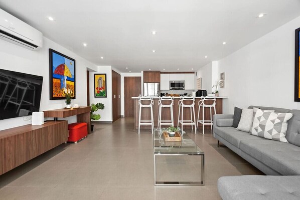A modern living room with a grey sofa, a coffee table, and a TV. There is a kitchen area with a dining set and a bar.