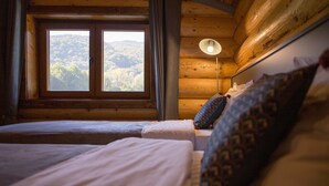 A wooden cabin room with two beds, a window with a view of the forest, and a bedside lamp.
