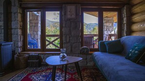 A cozy log cabin room with a sofa, a small table with a vase, and a view of the mountains through large windows.