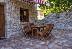 A stone building with a tiled patio, wooden table and chairs, and a striped awning.