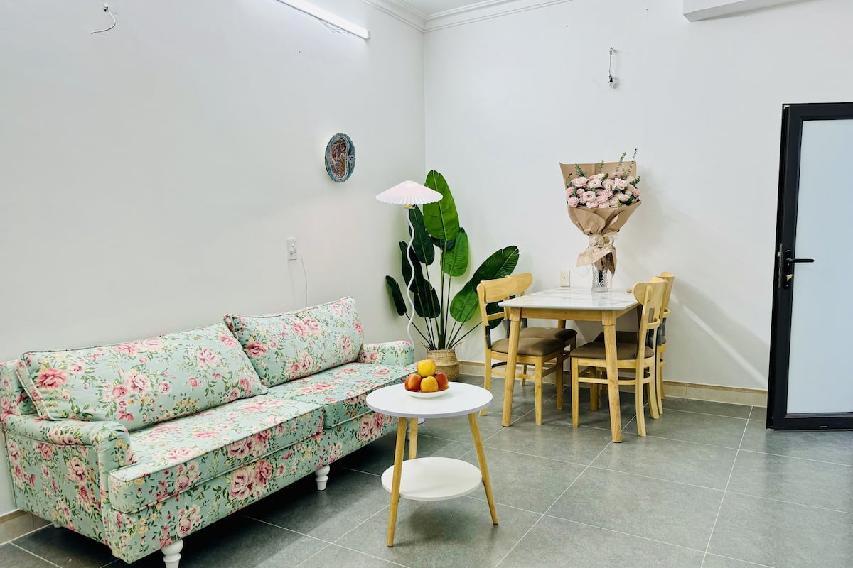 A living room with a floral sofa, a round table with chairs, a potted plant, and a bouquet of flowers.