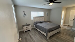 Une chambre à coucher comprenant un lit, des tables de chevet, un ventilateur de plafond, une fenêtre avec des stores et une porte.