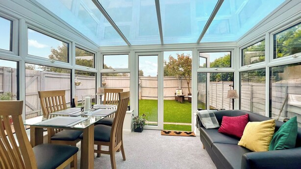 A conservatory with a glass roof, a dining table with chairs, and a sofa with cushions.