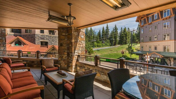 A covered outdoor seating area with wicker furniture and stone walls, overlooking a ski resort and buildings. - Catamount Lodge 214 - Northstar Village (Truckee)
