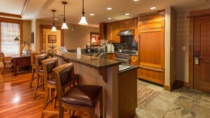 A modern kitchen with wooden cabinets, a central island, bar stools, and a dining area with a table and chairs. - Catamount Lodge 214 - Northstar Village (Truckee)
