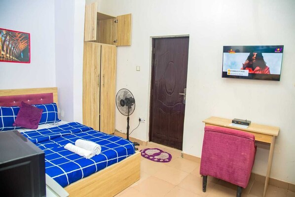 A bedroom with a bed, a TV, a wooden wardrobe, a fan, and a desk.