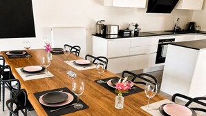 Un coin repas avec une longue table en bois dressée pour six personnes, des chaises noires et une cuisine en arrière-plan.