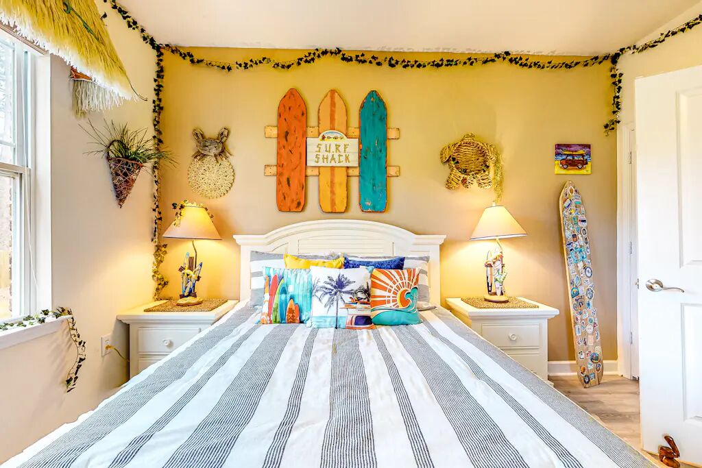 Een slaapkamer met een bed, twee nachtkastjes, een muur versierd met surfboards, en een surfboard dat tegen de muur staat, plus een raam.